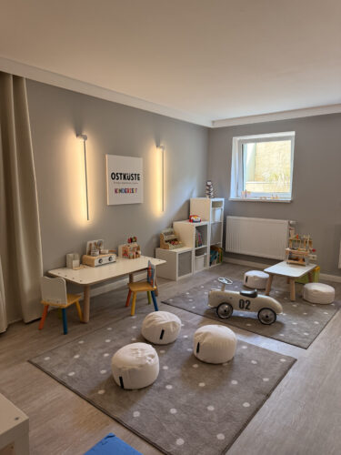 Großzügiges Spielzimmer mit grauen Teppichen mit weißen Punkten, Sitzkissen, Kinderküche, Holz-Rutschauto und Wandbild OSTKÜSTE Kinderzeit.