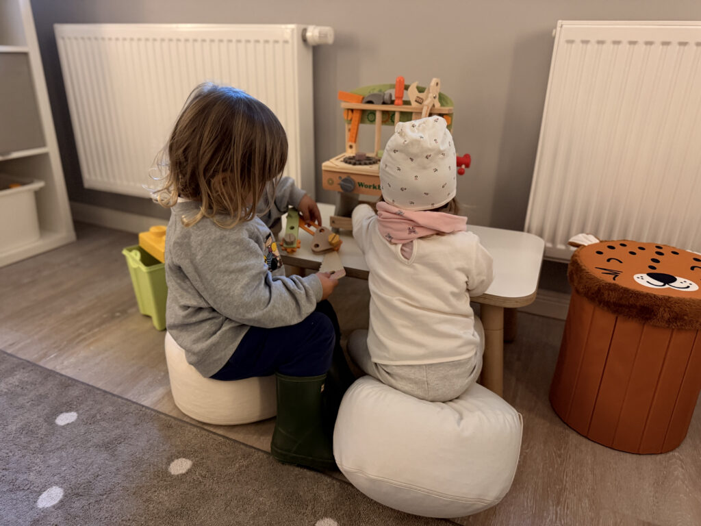 Zwei kleine Kinder sitzen auf weißen Hockern an einer Spiel-Werkbank aus Holz im Spielzimmer.