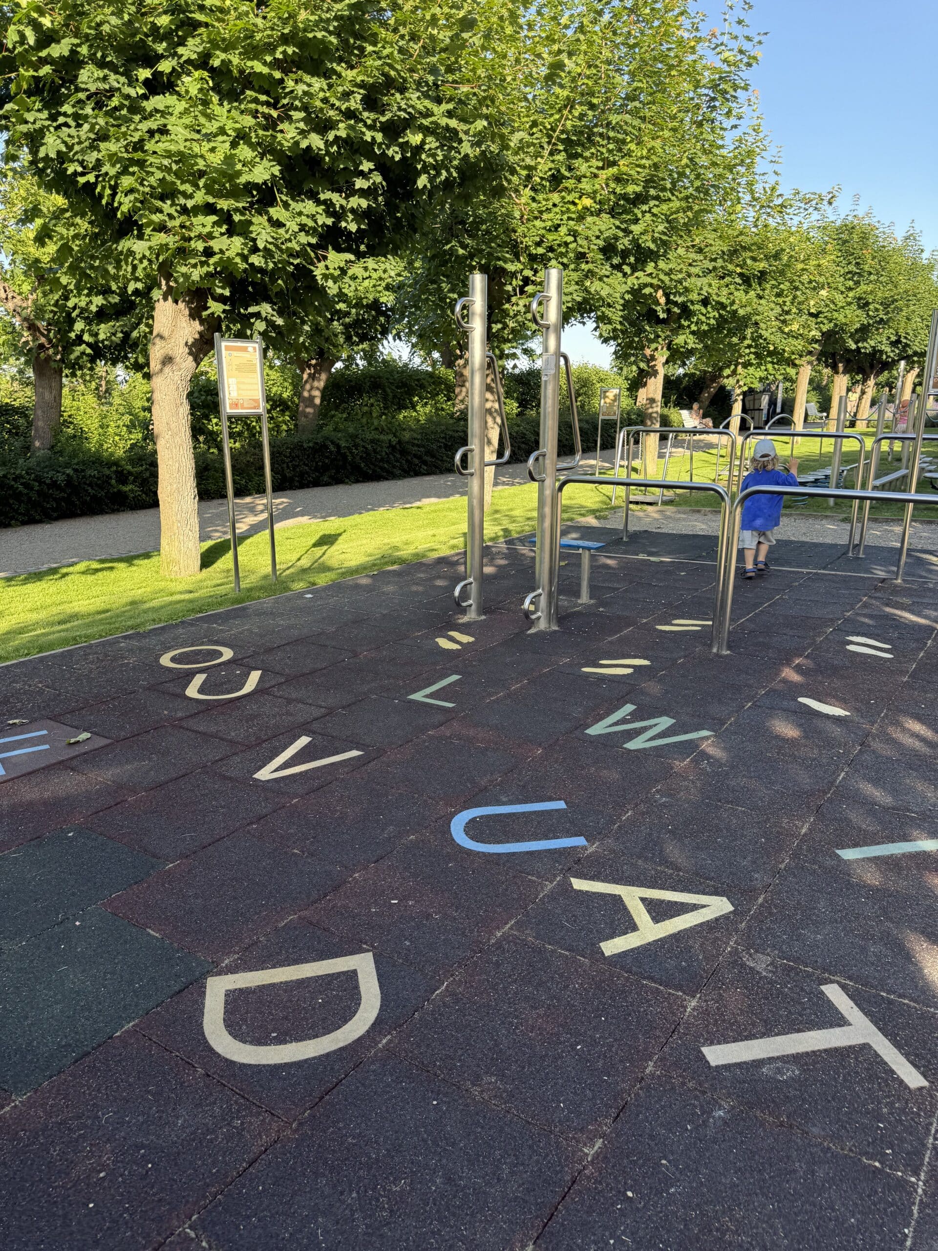 Aktivspielplatz mit Buchstabe-Aufdrucken auf dem Fallschutzboden und Edelstahl-Sportgeräten unter einer baumbestandenen Promenade.