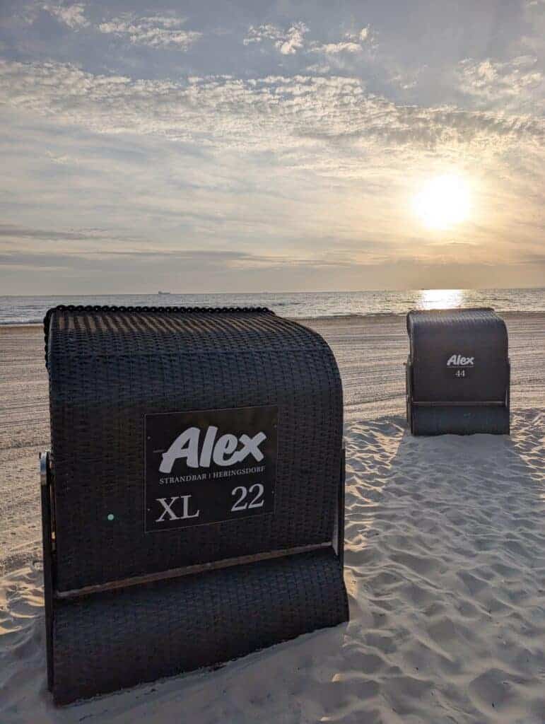 Zwei dunkle Strandkörbe mit Aufschrift der Alex Strandbar stehen im Sand vor der ruhigen Ostsee bei untergehender Sonne.