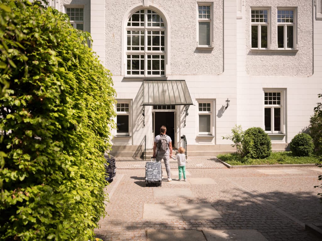 Ein Mann mit Rucksack und Koffer geht Hand in Hand mit einem kleinen Kind auf den eleganten Eingang der weißen, historischen Villa Staudt zu.