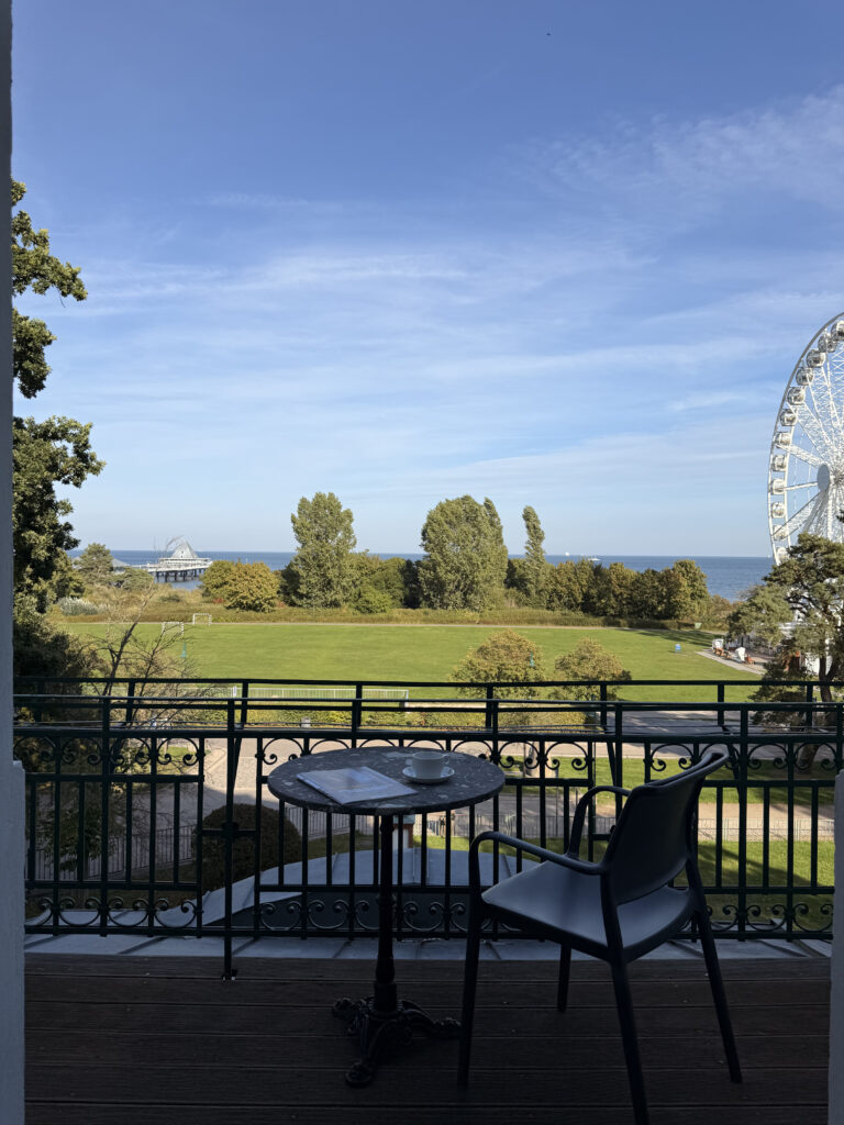 Dunkler Balkon mit rundem Tisch, Kaffeetasse und Stuhl. Im Hintergrund erstreckt sich eine grüne Wiese vor der Ostsee mit Seebrücke und Riesenrad.