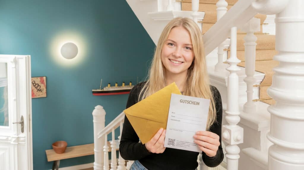 Eine lächelnde Frau steht im lichtdurchfluteten Treppenhaus einer Villa und präsentiert den hochwertigen OSTKÜSTE Gutschein samt goldenem Umschlag.