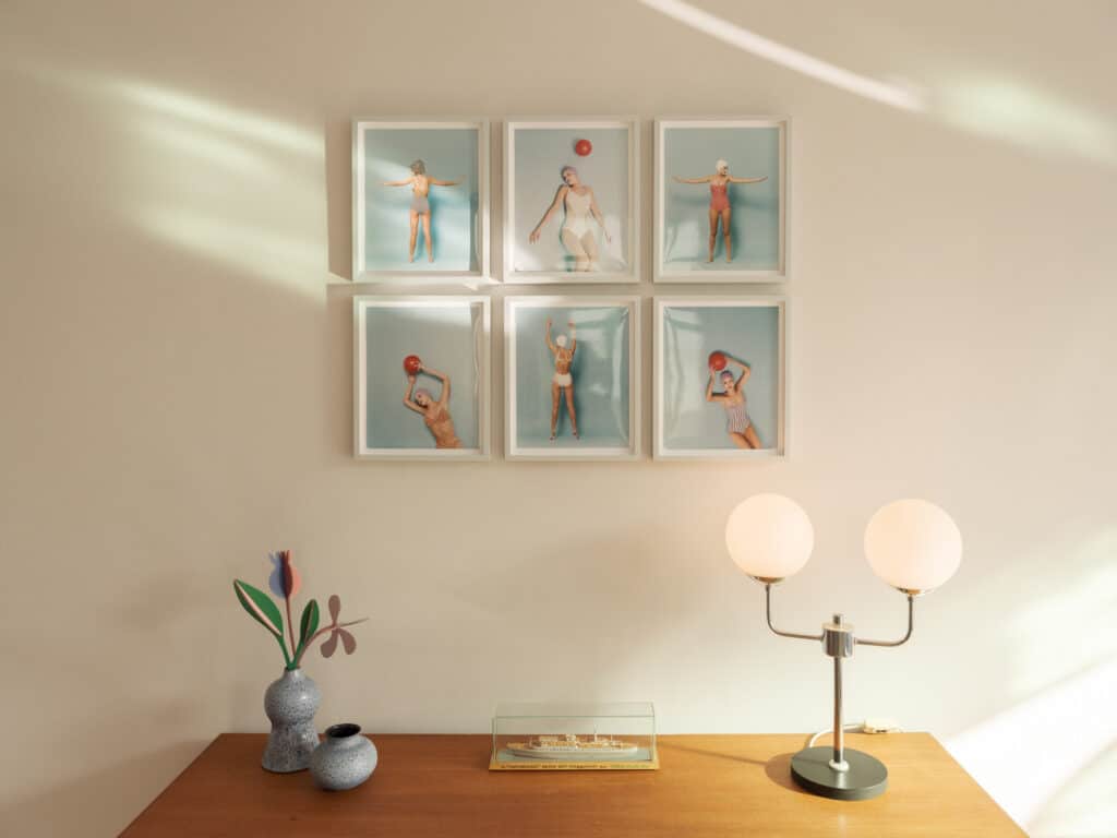 Sechs gerahmte Retro-Bilder mit Schwimmer-Motiven an einer hellen Wand über einem hölzernen Sideboard mit einer Tischleuchte und Vasen.