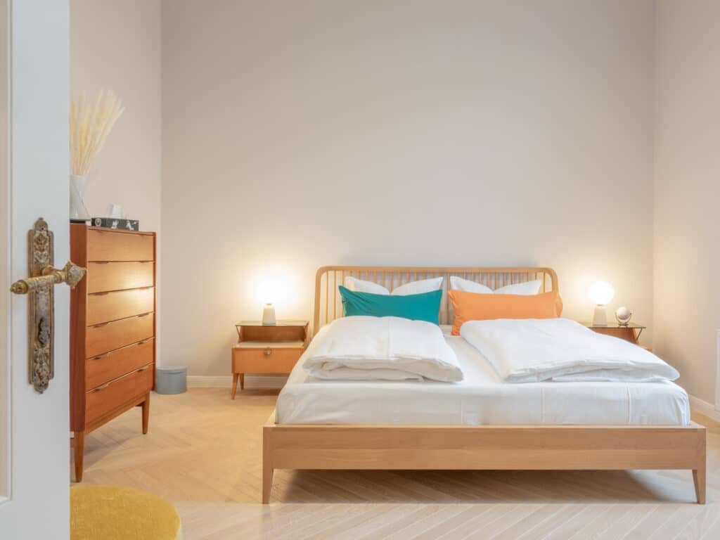 Helles Schlafzimmer mit Doppelbett aus Holz, weißer Bettwäsche, bunten Kissen, Mid-Century-Kommode und historischem Türbeschlag im Vordergrund.