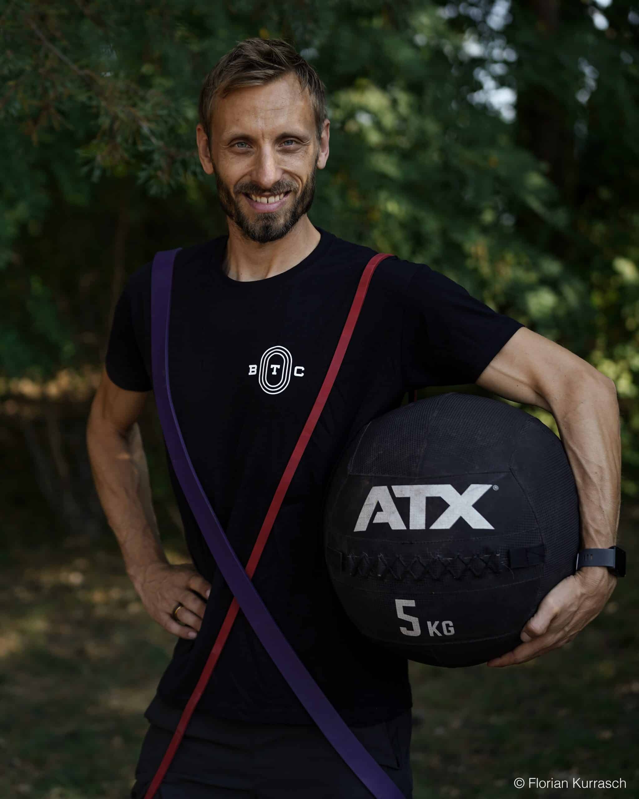 Porträt des lächelnden Lauftrainers Benjamin Schalanda in einem schwarzen BTC-Sportshirt, der ein rotes und violettes Fitnessband über den Schultern trägt und eine schwarze Kugelhantel hält.