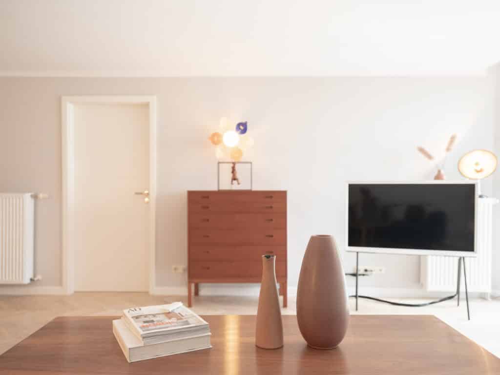 Braune Holzkommode mit einer dekorativen bunten Lampe; daneben steht ein weiß eingerahmter Fernseher auf einem schwarzen Stativ.