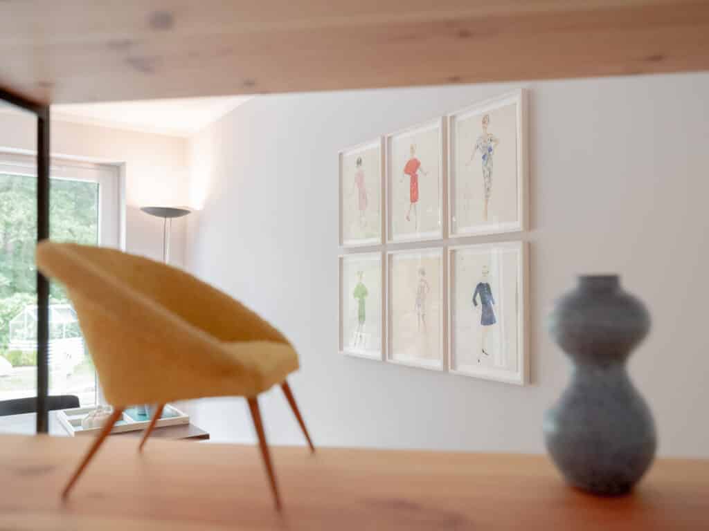 Ein brauner Sessel mit geflochtener Lederbespannung steht zentral vor einer weißen Wand mit gerahmten Skizzen und einer weißen Vase.