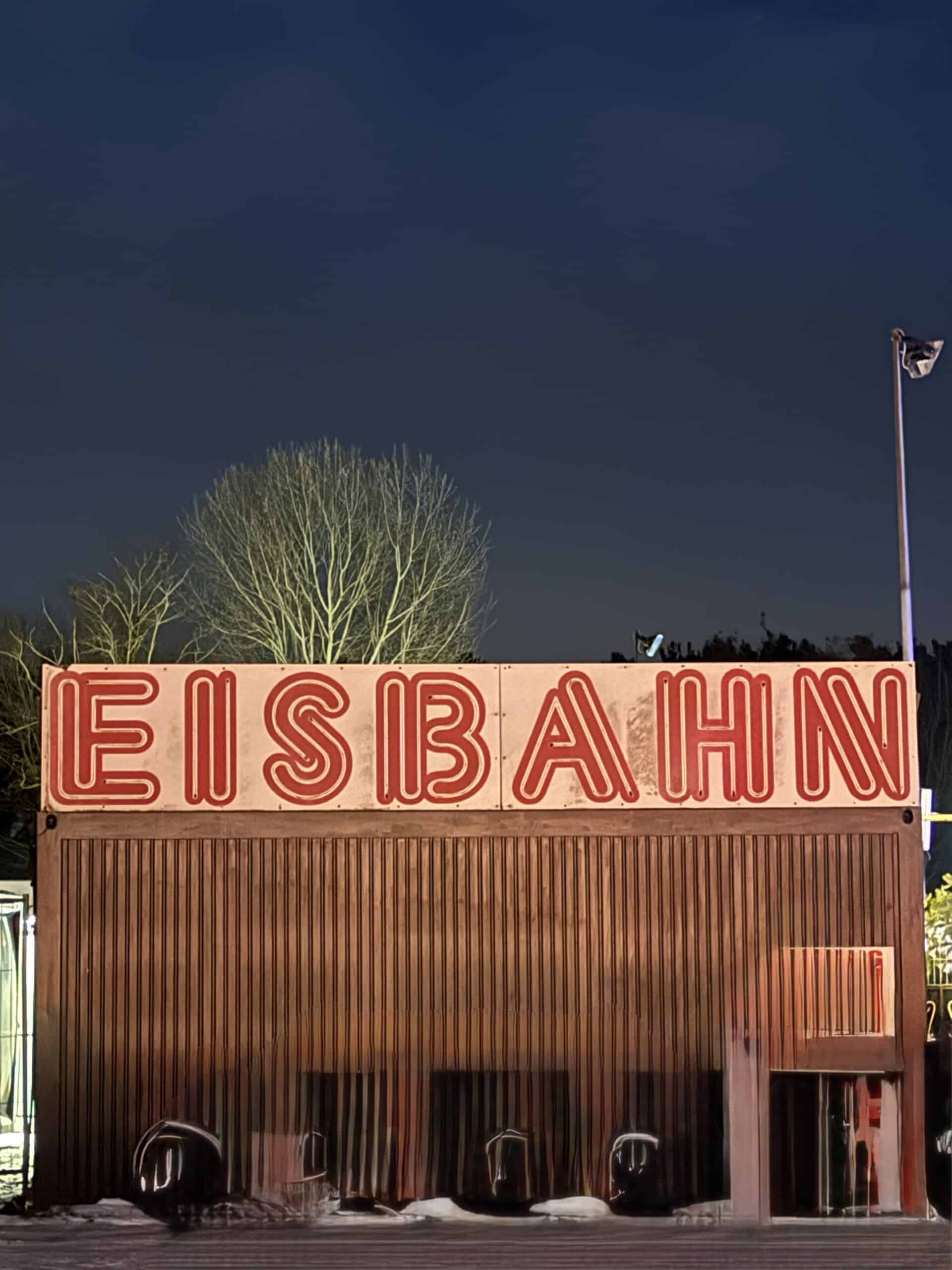 Leuchtender roter Neonschriftzug Eisbahn auf einem braunen Holzcontainer vor dunkelblauem Abendhimmel.
