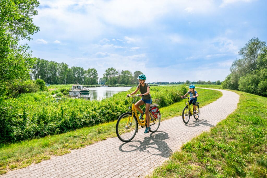 Zwei Kinder fahren auf gelben Fahrrädern auf einem gepflasterten Radweg an einem Deich neben einem bewachsenen Ufer mit Booten.