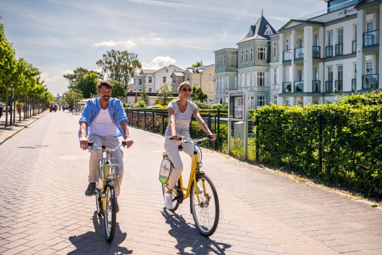 Ein lächelndes Paar radelt auf gelben Fahrrädern nebeneinander über eine sonnige Promenade entlang historischer Bäderarchitektur-Villen.