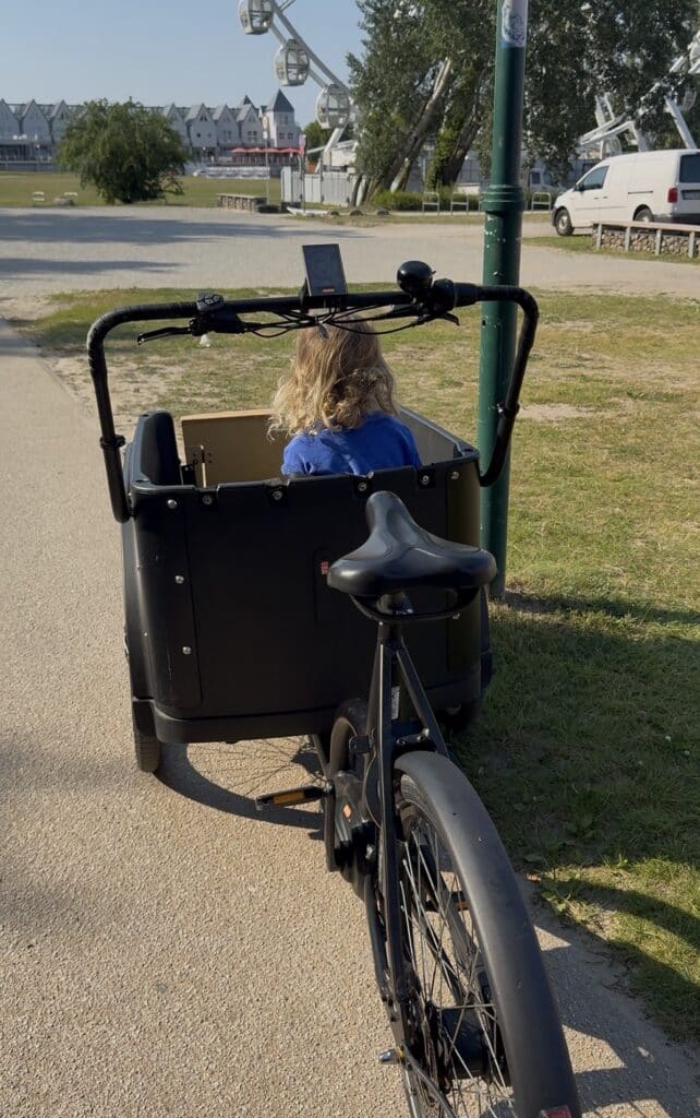 Perspektive vom Sattel eines schwarzen E-Lastenrads mit Kind in der Transportbox auf einen asphaltierten Küstenweg nahe einem Riesenrad.