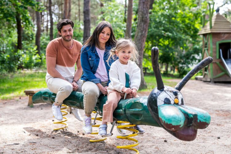 Vater, Mutter und kleine Tochter sitzen gemeinsam lachend auf einer grünen Feder-Raupe auf einem Waldspielplatz im Sonnenlicht.