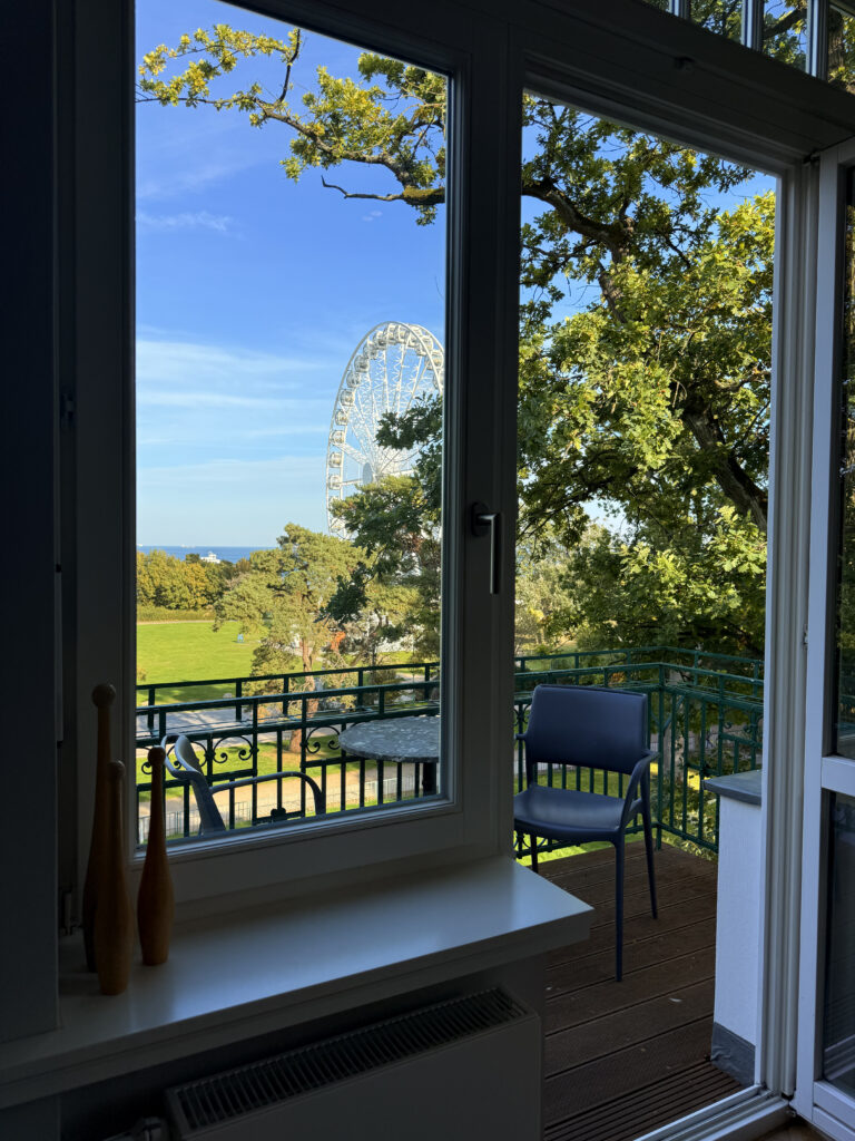 Blick durch ein geöffnetes weißes Fenster auf einen Balkon, über grüne Baumkronen hinweg auf ein weißes Riesenrad und die blaue Ostsee.