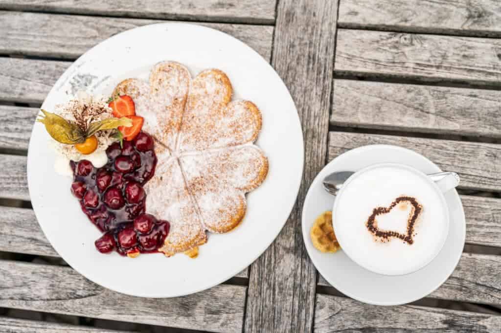 Flacher Teller aus der Vogelperspektive mit einer herzförmigen Waffel, Kirschgrütze, Beeren und Sahne neben einer Tasse Cappuccino mit Schaumherz.