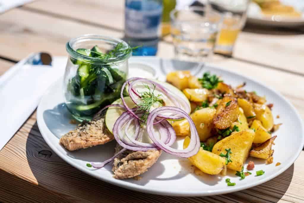 Ein weißer Teller mit gebratenem Fischfilet, Apfelscheiben, roten Zwiebeln, knusprigen Bratkartoffeln und einem Einmachglas voller Gurkensalat.
