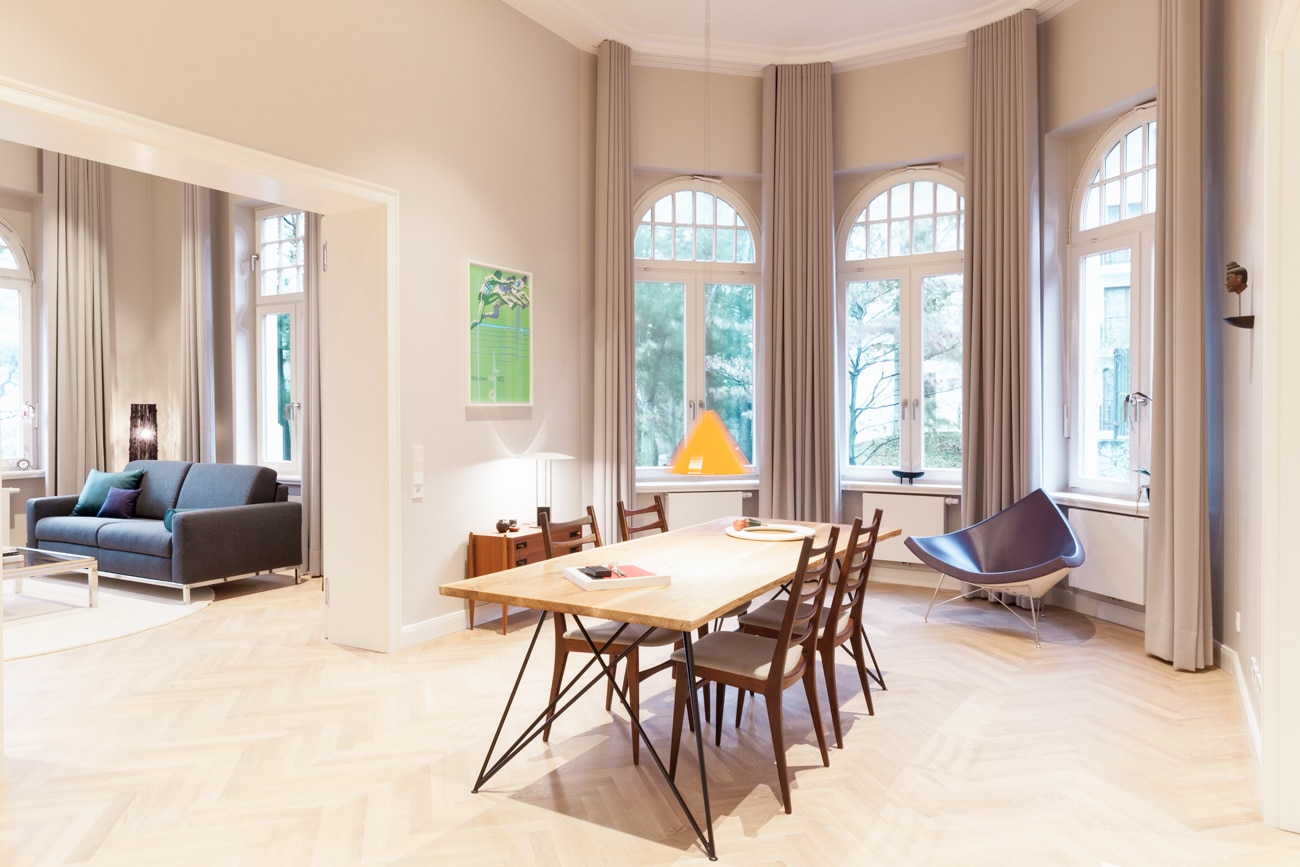 Ein prachtvoller, hoher Raum mit Fischgrätparkett und großen Rundbogenfenstern, in dessen Mitte ein langer Esstisch aus Holz mit sechs Design-Stühlen steht.