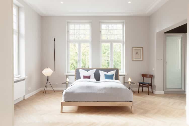 Großzügiges Schlafzimmer mit einem Doppelbett aus Holz, hohen Sprossenfenstern zum Garten, Fischgrätparkett und stilvollen Designlampen.