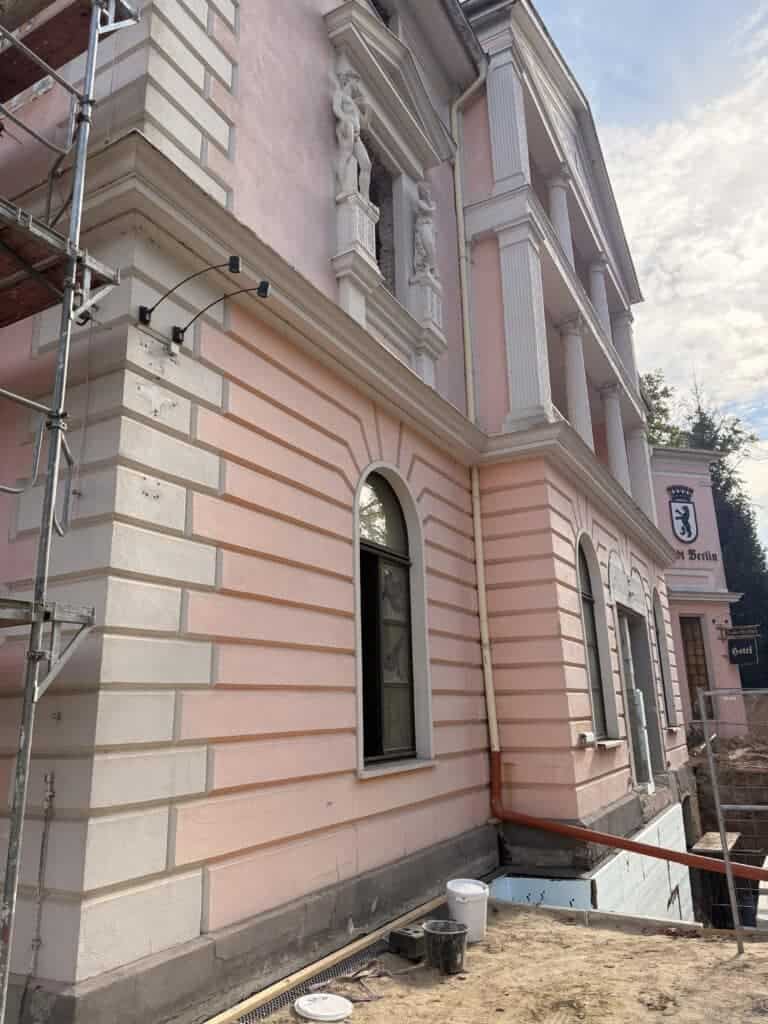 Detailansicht der rosa Stuckfassade mit verzierten Bogenfenstern, weißen Säulen, Skulpturen und Baugerüst an der Gebäudeecke.