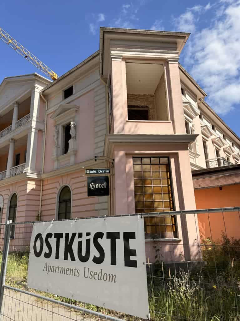Rosa-weiße Fassade eines historischen Bäderarchitektur-Gebäudes mit Bauzaun und großem OSTKÜSTE-Banner im Vordergrund.