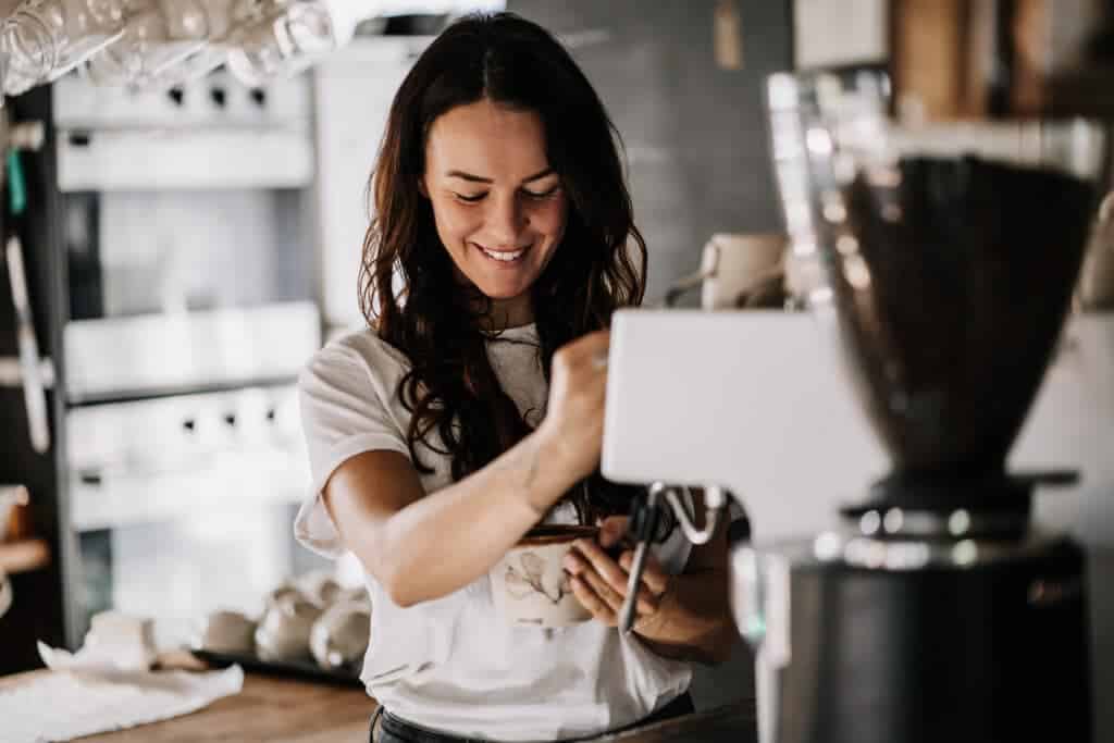 Eine dunkelhaarige Frau im weißen T-Shirt lächelt herzlich, während sie an einer Siebträgermaschine eine Keramiktasse zubereitet.