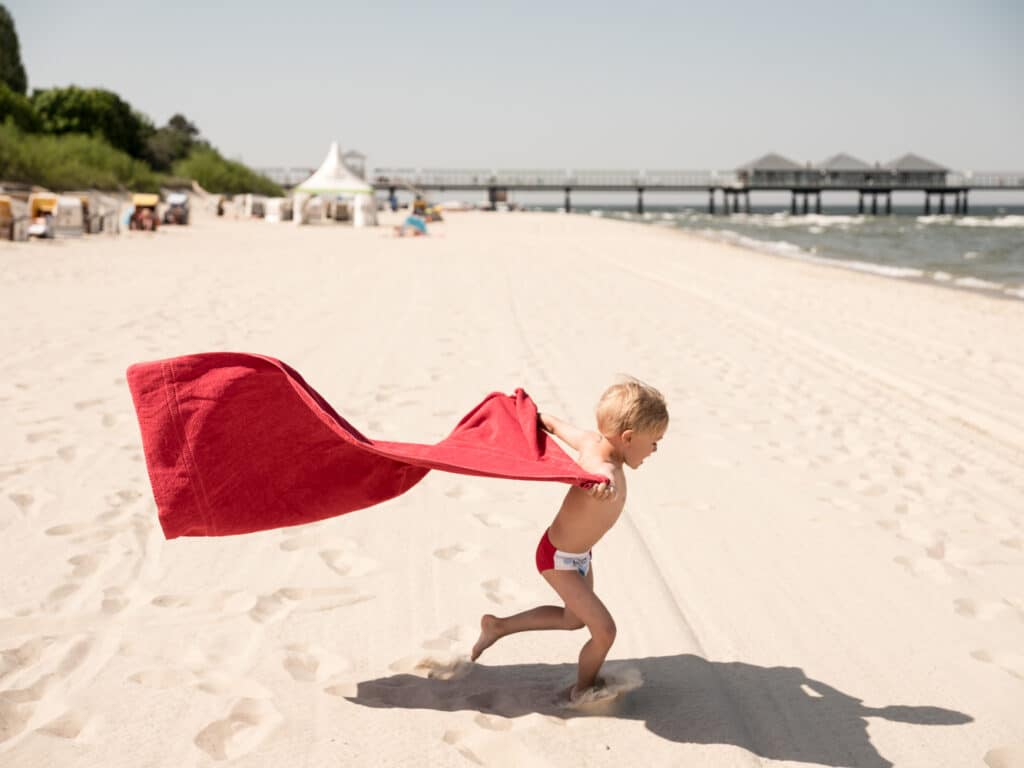 Ein kleines Kind in roter Badehose rennt mit einem großen, im Wind wehenden roten Badetuch über den breiten Sandstrand; im Hintergrund ist die Ahlbecker Seebrücke im Meer zu sehen.