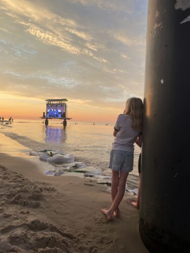 Ein junges Mädchen mit blonden Haaren steht abends am Strand und blickt auf eine beleuchtete Musikbühne, die auf Pfählen im ruhigen Wasser vor einem farbenfrohen Abendhimmel steht.