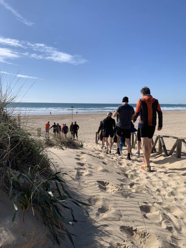 Rückansicht einer sportlichen Gruppe in Laufkleidung, die über einen sandigen Dünenweg mit Holzgeländer hinab zum breiten Sandstrand und der Ostsee geht.