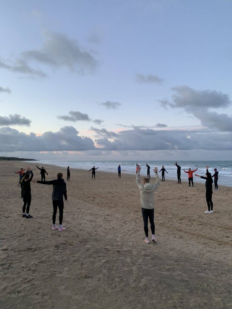 Eine Gruppe von Menschen steht im Kreis auf dem weiten Sandstrand und führt unter bewölktem Morgenhimmel Dehnübungen mit ausgestreckten Armen aus.