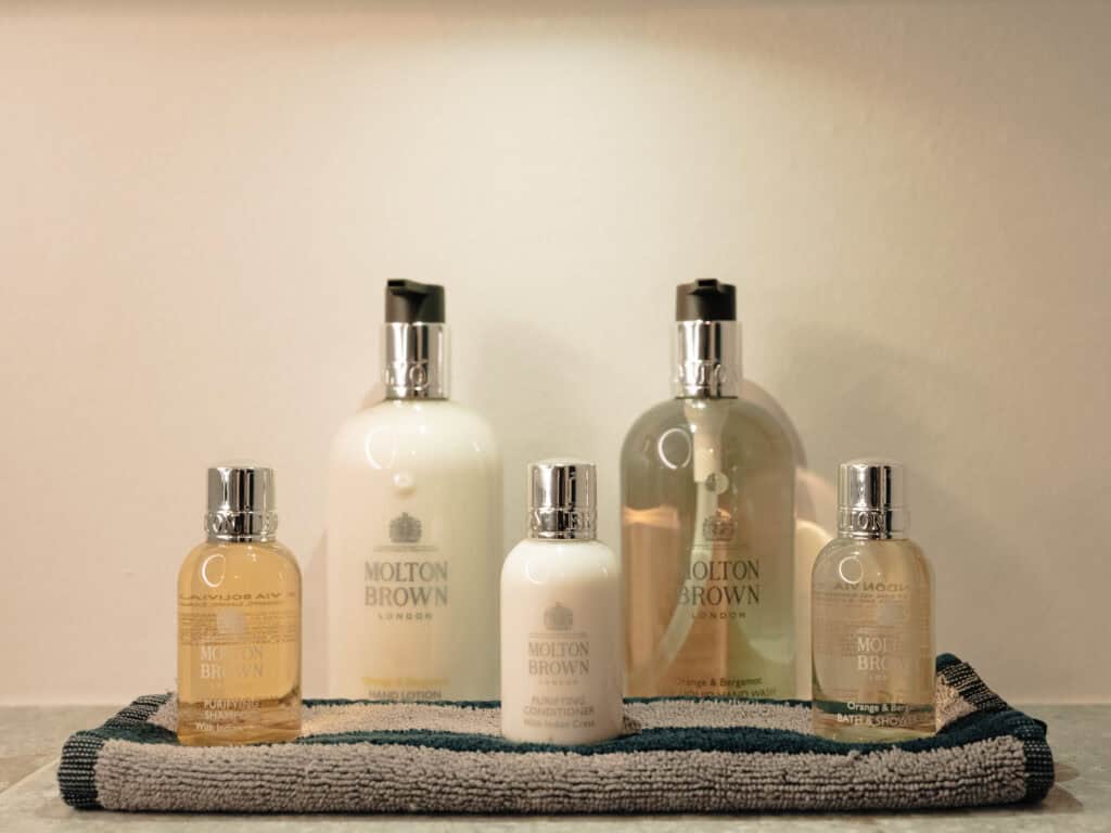 Fünf Flakons mit Pflegeprodukten von Molton Brown London stehen aufgereiht auf einem gestreiften Frottee-Gästehandtuch.