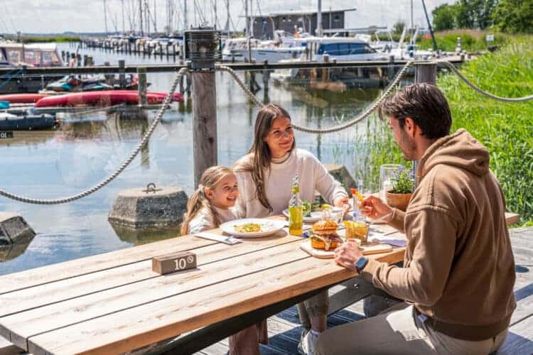 Eine dreiköpfige Familie sitzt an einem rustikalen Holztisch im Außenbereich und speist mit Blick auf Segelboote in einem sonnigen Hafen.