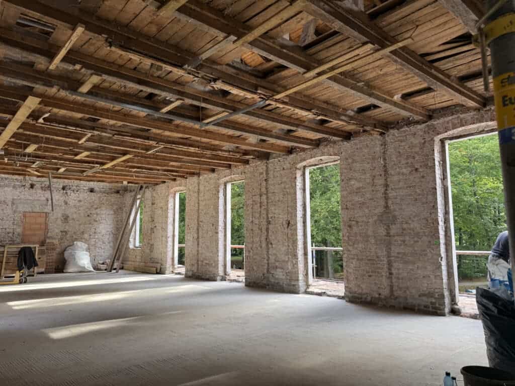 Weitläufiger Innenraum im Rohbau mit freigelegter Holzbalkendecke, Ziegelmauerwerk und großen Fensteröffnungen mit Blick ins Grüne.