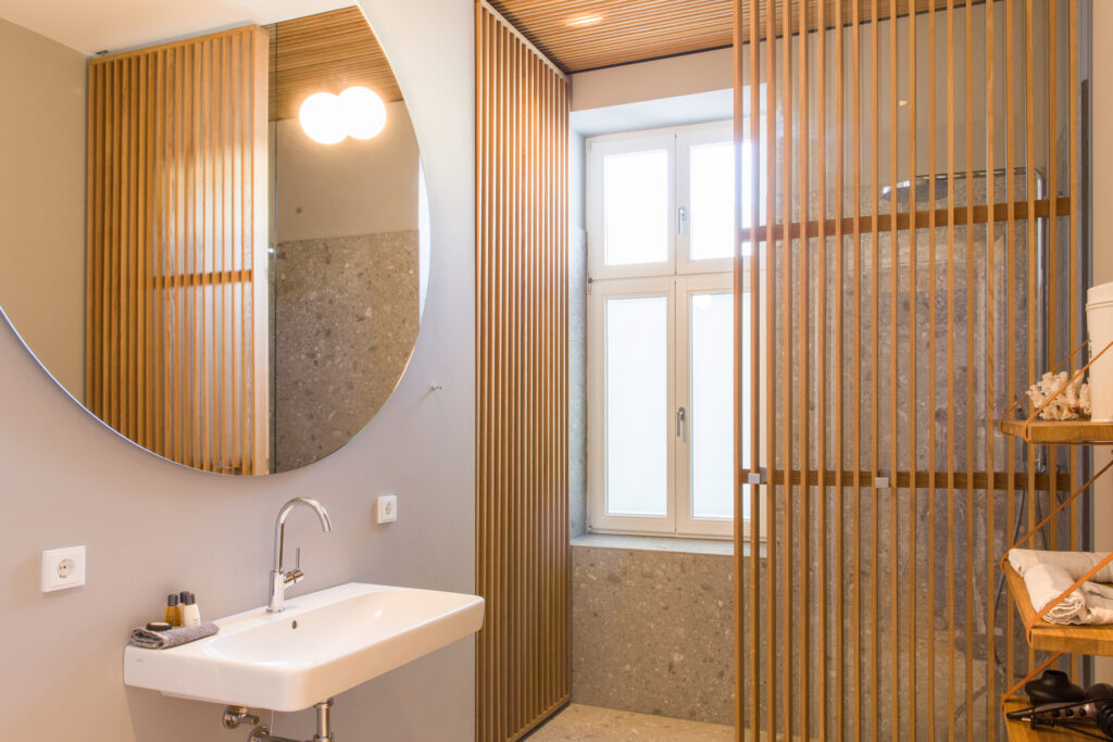 Moderner Waschplatz mit weißem Waschbecken, rundem Spiegel und vertikalen Holzlamellen als Abtrennung zum Duschbereich mit hellen Fliesen.