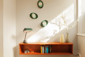 Wandmontiertes Holzregal mit Büchern, einer grünen Schreibtischlampe, Keramikvasen mit Zweigen und kreisförmiger Wanddekoration darüber.