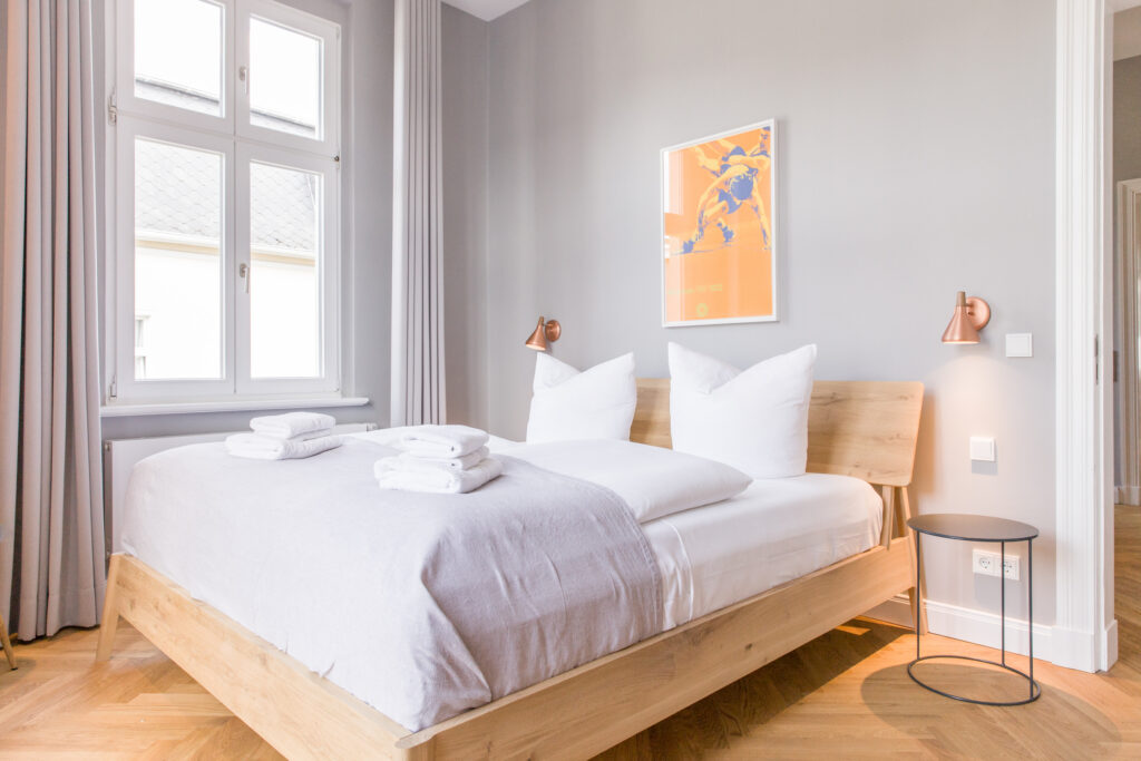 Großes hölzernes Doppelbett mit weißer Bettwäsche und Handtuchstapeln auf hellem Fischgrätparkett neben hohem Fenster.