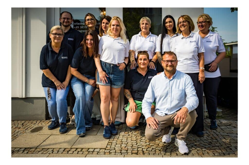 Elf lächelnde Mitarbeiterinnen und Mitarbeiter des OSTKÜSTE Teams stehen gemeinsam vor dem Gebäude des Gästeempfangs in Ahlbeck.
