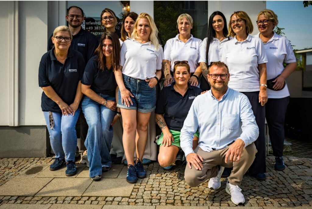Elf lächelnde Mitarbeiterinnen und Mitarbeiter des OSTKÜSTE Teams stehen gemeinsam vor dem Gebäude des Gästeempfangs in Ahlbeck.