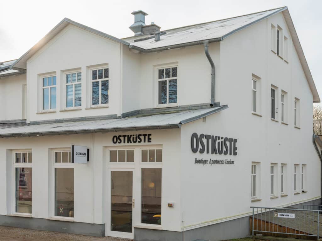 Außenansicht eines zweistöckigen weißen Gebäudes mit dem Logo der Ostküste Boutique Apartments und einem Parkplatzschild.