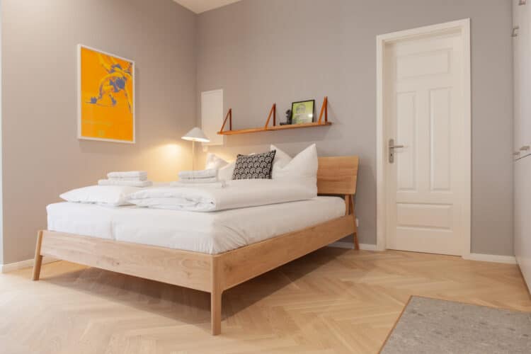 Heller Schlafbereich mit großem Doppelbett aus hellem Holz, Fischgrätparkett, weißer Bettwäsche und modernem gelben Kunstwerk an der Wand.