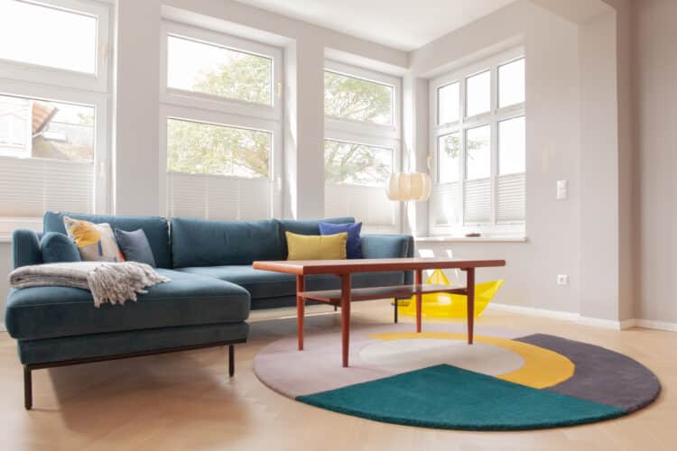 Lichtdurchfluteter Wohnbereich mit großem, blauen Ecksofa, Couchtisch aus Holz und rundem Design-Teppich vor einer Fensterfront.