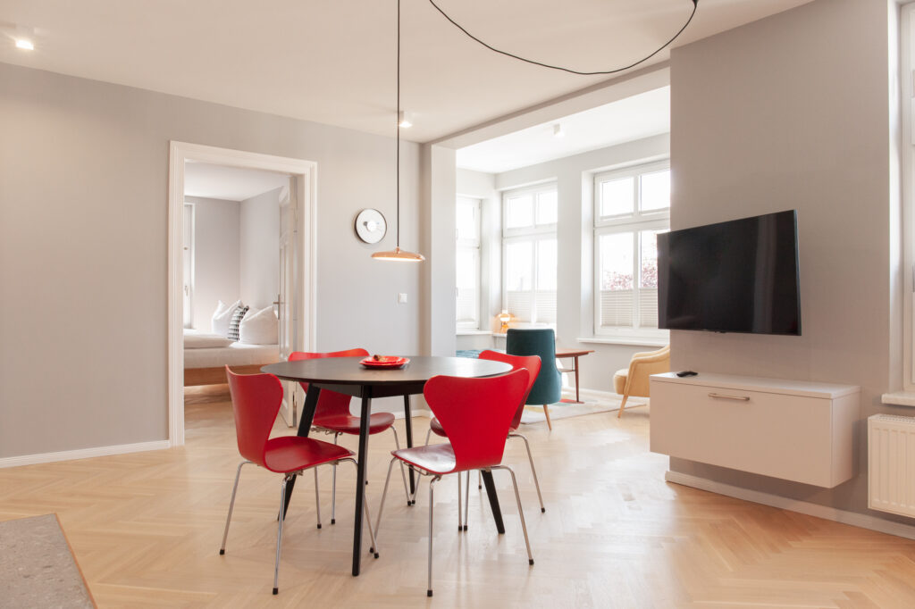 Offener Wohn-Essbereich mit rundem Tisch und roten Design-Stühlen, TV an der Wand und Blick durch eine Tür ins separate Schlafzimmer.