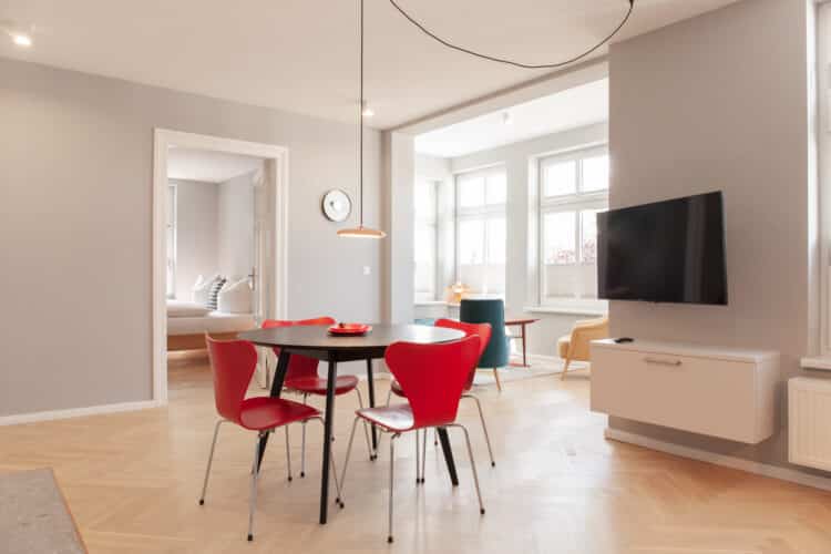 Offener Wohn-Essbereich mit rundem Tisch und roten Design-Stühlen, TV an der Wand und Blick durch eine Tür ins separate Schlafzimmer.