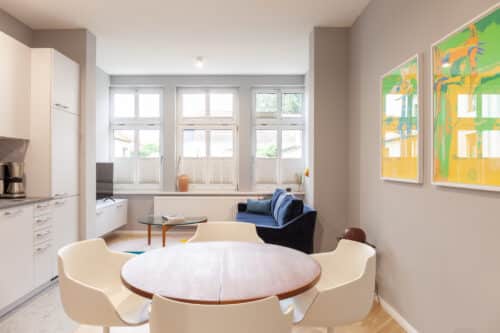 Heller, offener Wohn-Essbereich im Apartment 5 mit rundem Holztisch, weißen Design-Stühlen, blauem Sofa und "München 1972" Kunst an der Wand.