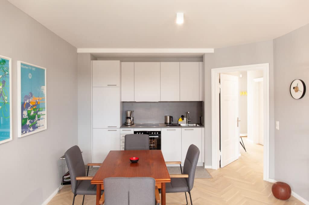 Heller, offener Essbereich mit Holztisch und grauen Stühlen vor einer weißen Küchenzeile, "München 1972" Kunst an der Wand.