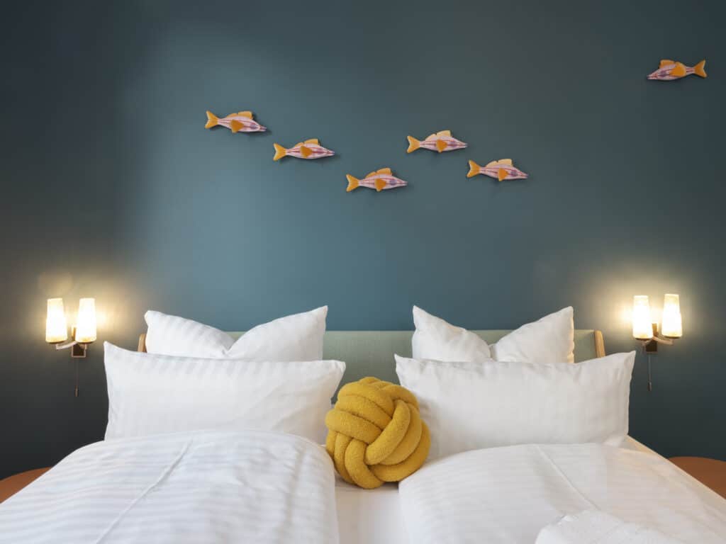 Doppelbett mit weißen Kissen und Decken, verziert mit einem gelben Knotenkissen vor einer petrolblauen Wand mit Deko-Holzfischen[cite: 4, 5].
