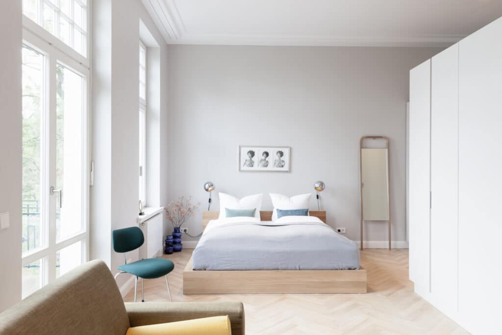 Hell gestaltetes Schlafzimmer mit einem Doppelbett aus Holz, edlem Fischgrätparkett, einem dunkeltürkisen Stuhl und großen Fenstern.