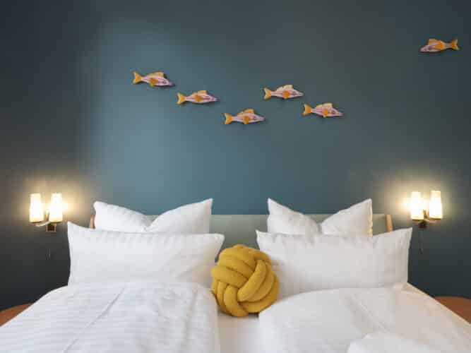Ein Doppelbett mit weißer Bettwäsche und einem markanten senfgelben Knotenkissen, das vor einer dunkelblauen Wand steht, an der sechs hölzerne Deko-Fische befestigt sind.