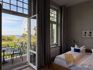 Helles Schlafzimmer mit Doppelbett und geöffneter Balkontür, die den Blick auf einen Balkon, ein Riesenrad und die Ostsee freigibt.