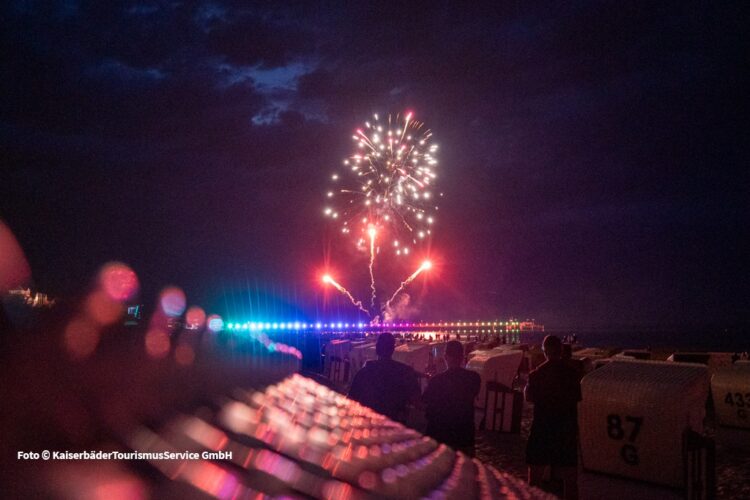Funkelndes Feuerwerk steilt über einer bunte beleuchteten Seebrücke in der Dämmerung auf, während Menschen im Vordergrund zusehen.