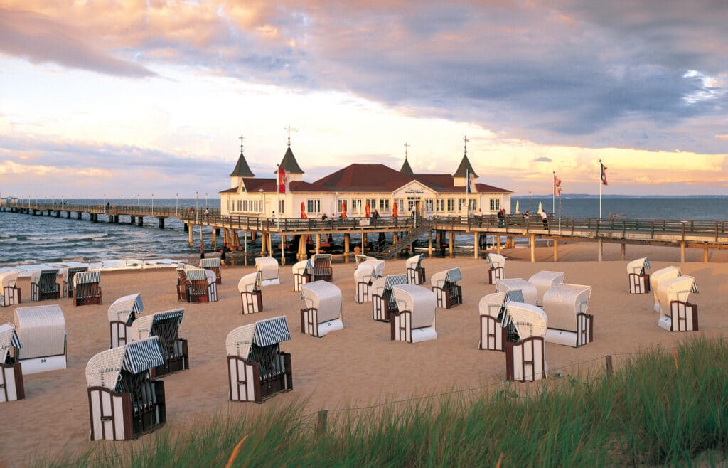 Historische weiße Seebrücke Ahlbeck mit Türmchen am Ostseestrand bei Abenddämmerung, davor stehen viele Strandkörbe geordnet im Sand.